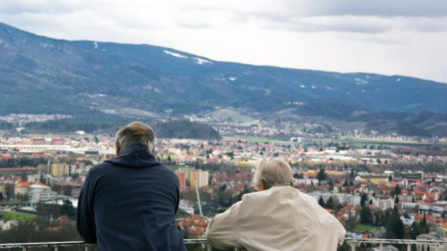 Couple of old seniors look around from lookout tower on city and hills in the background.