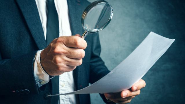 Tax inspector investigating financial documents through magnifying glass, forensic accounting or financial forensics, inspecting offshore company financial papers, documents and reports.