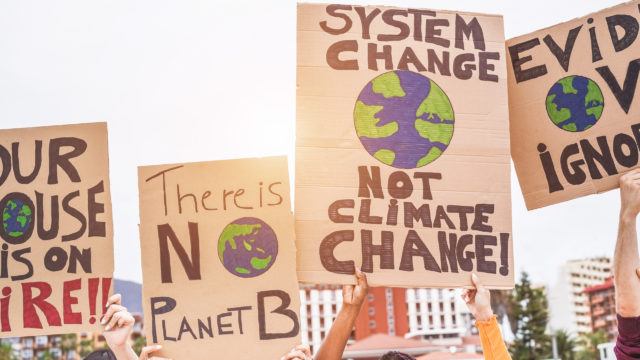 a group of climate change protestors with signs that read “our house is on fire”, “there is no planet b”, “system change not climate change” and “evidence ignored”