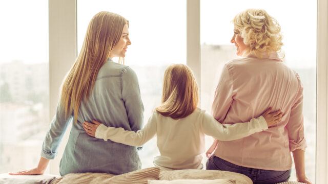 Back view of three generations of women sitting on sofa in front of a window.