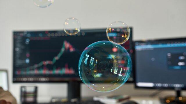 Is there an AI bubble? Is it about to burst? Your questions answered Soap bubbles floating over a desk with a computer screen showing stock market charts in the background.