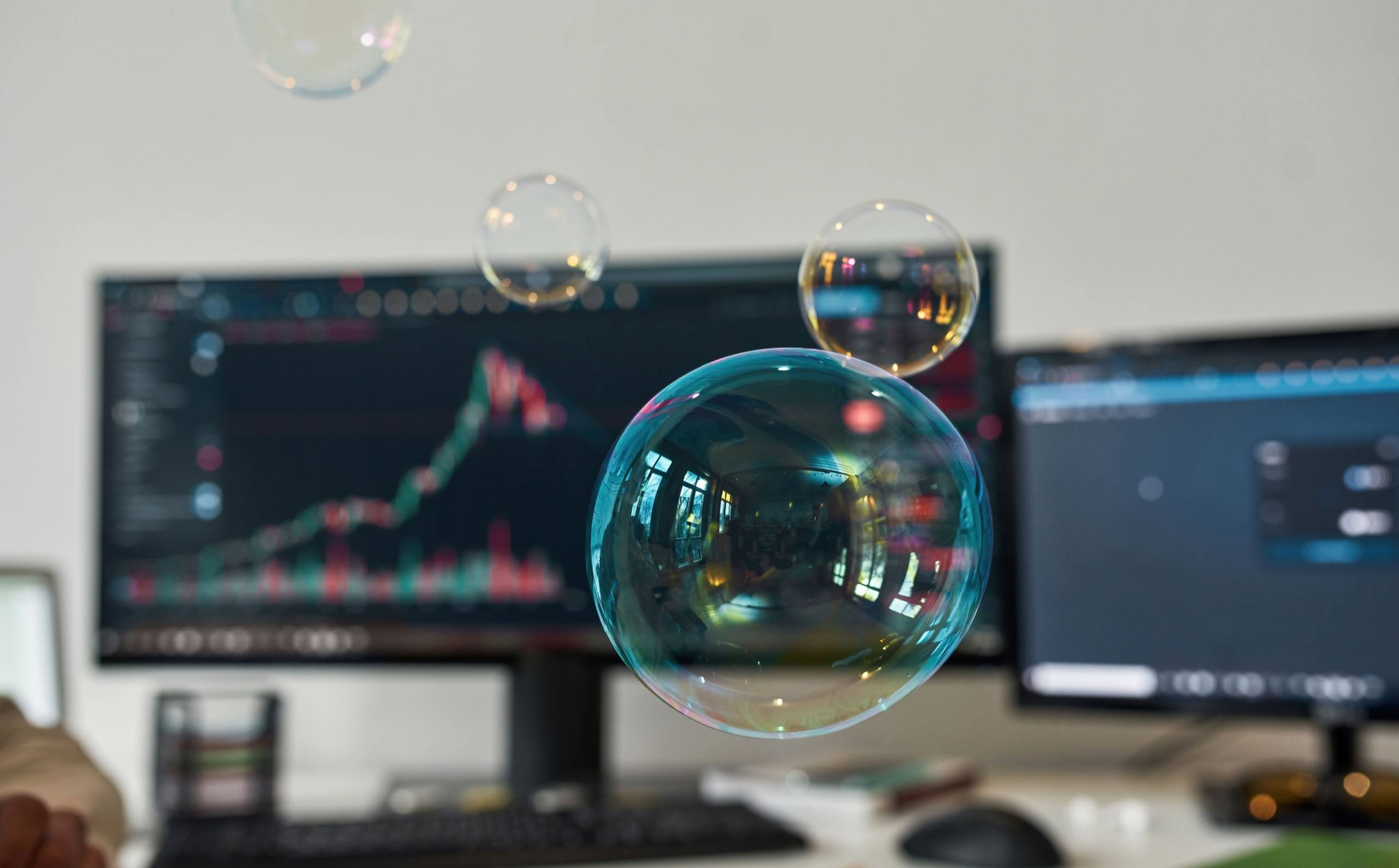 Home Soap bubbles floating over a desk with a computer screen showing stock market charts in the background.